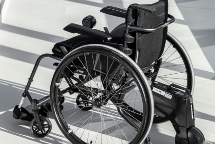 Mūsdienīgs melns ratiņkrēsls ar lieliem aizmugurējiem riteņiem, maziem priekšējiem riteņiem un pievienotu elektrisko piedziņu atrodas uz gaišas grīdas saules apspīdētā telpā.