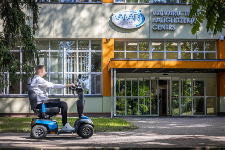 Vīrietis brauc ar zilu skrejriteni pie modernas ēkas, virs kuras ieejas ir uzraksts Vaivaru Palīgierīču centrs. Ēkai ir lieli logi un blakus aug koks.