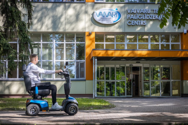 Vīrietis brauc ar zilu skrejriteni pie modernas ēkas, virs kuras ieejas ir uzraksts Vaivaru Palīgierīču centrs. Ēkai ir lieli logi un blakus aug koks.