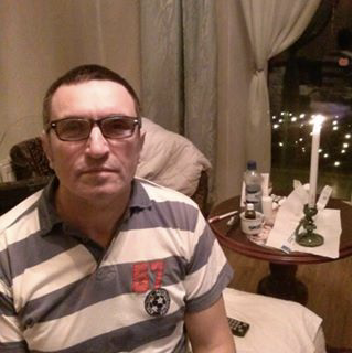 Vīrietis ar brillēm un svītrainā polo kreklā sēž iekštelpās. Aiz viņa uz apaļa galda ir novietota aizdegta svece, ūdens pudele un dažādi nelieli priekšmeti. Aiz loga redzamas pasaku gaismas.