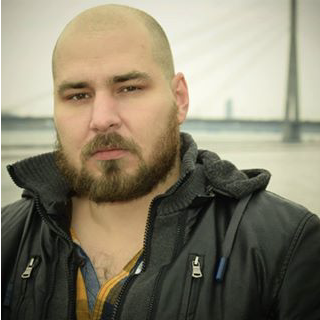 Bārdains vīrietis ar skūtu galvu melnā jakā stāv ārā pie ūdens, neskaidrā fonā redzams tilts un kabeļi.
